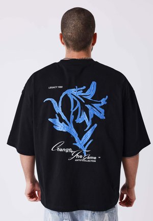 Personne aux cheveux courts porte un T-shirt noir oversize avec un motif abstrait de feuille bleue et un texte blanc "Change The Game" dans le dos.
