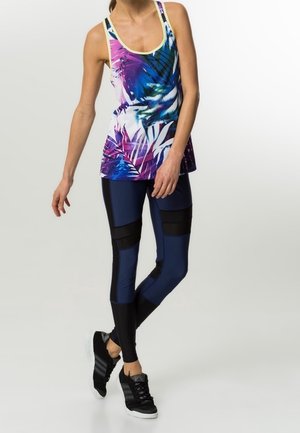 Donna che indossa un top smanicato a stampa tropicale viola, blu e bianco con leggings blu navy e neri e scarpe sportive nere, in piedi con una gamba incrociata.