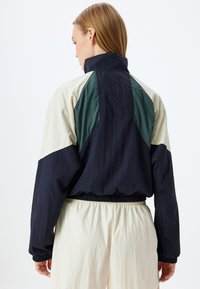 Tuulitakki, jossa on tummansinisiä, vihreitä ja kermahenkisiä paneeleja, joustavat hihansuut ja helma; kevyt, rakennetekstiili ja korkea kaulus.