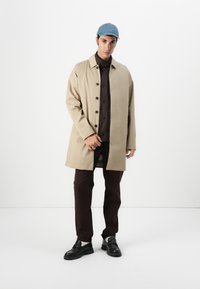 Uma pessoa está de pé contra um fundo branco, vestindo um casaco bege, uma camisa castanha escura e calças, complementados por um gorro azul e sapatos pretos.