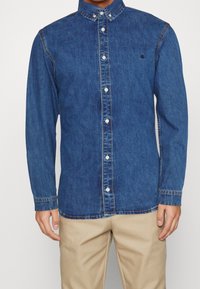 Jeansskjorta i mörkblått med långa ärmar, knapp-down krage, framknappar och en subtil logotyp på bröstet. Slät textur.