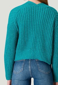 Türkiser Strickpullover mit strukturiertem Muster, verkürztem Schnitt und geripptem Saum, kombiniert mit blauen Jeans im Fünf-Taschen-Stil.