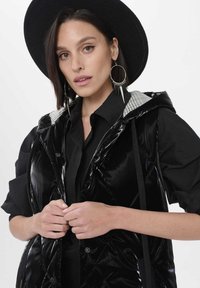 Czarna, błyszcząca kamizelka puchowa z kapturem, w romboidalny wzór pikowania i w kratę podszewka. Nosi ją osoba w czarnej koszuli, z dużymi kolczykami w kształcie kół.