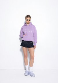 Liliowy sweatshirt z kapturem z srebrnymi literami, w połączeniu z ciemnoniebieską spódniczką mini, białymi skarpetkami do kolan i jasnoniebieskimi klapkami Crocs.