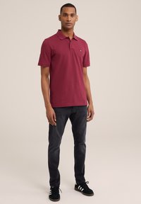 Burgundijos polo marškiniai su trumpomis rankovėmis ir apykakle, derinami su tamsiomis džinsinėmis kelnėmis ir juodais sportbačiais su baltais dryžiais. Paprastas logotipas krūtinėje.