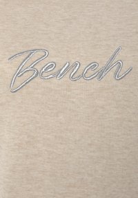 Felpa in cotone beige con logo "Bench" ricamato in argento in corsivo. Texture morbida con un motivo leggermente melangiato.