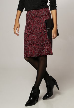 Jupe longueur genou à motif floral rouge avec une texture lisse, associée à des collants opaques noirs et des bottines, tenant une pochette noire.