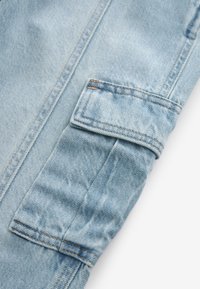 Tissu en denim bleu clair avec une poche à rabat rectangulaire présentant des coutures visibles et de légers plis sur le tissu.