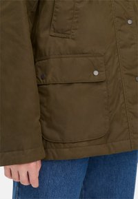 Olivengroen jack met een gladde textuur, voorzien van een klepzak en zilveren drukknoopjes aan de voorkant. Draagt over een blauwe spijkerbroek.
