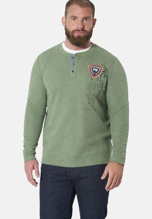 Man with beard wearing green long-sleeve henley shirt with emblem and printed text, paired with dark blue jeans, standing against plain background.
