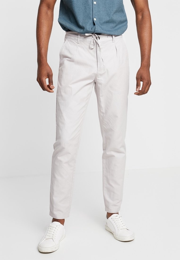 Chinos gris clair avec une taille élastique à cordon de serrage, coupe effilée, et poches latérales, accompagnés de baskets blanches sur un fond uni.