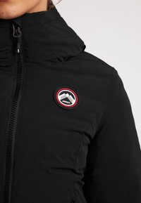 Sort isoleret jakke med højt krave, frontlynlås og et rundt logo-patch i rød og sort med bjergdesign på øverste venstre side.