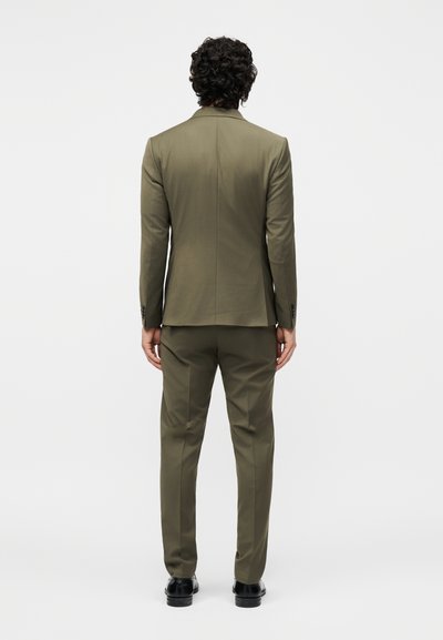 Άνδρας με σγουρά μαύρα μαλλιά που φοράει ένα πράσινο-ελιά κοστούμι και μαύρα παπούτσια, στέκεται όρθιος με την πλάτη προς τα εμπρός σε μια μονόχρωμη φόντο.