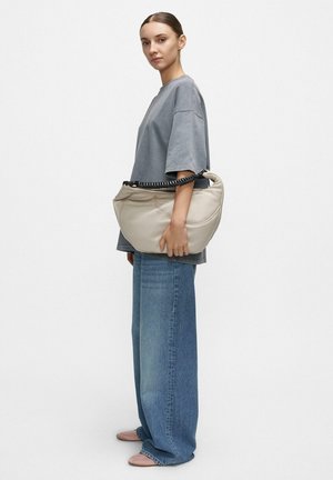 Young woman stands sideways wearing loose gray T-shirt and wide blue jeans, holding beige handbag with black chain strap.