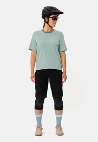 T-shirt à manches courtes vert clair évacuant l'humidité, shorts noirs, genouillères, chaussettes bleu clair à rayures et chaussures beiges. Casque noir et lunettes de soleil.