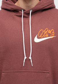 Gros plan d'une personne portant un sweat à capuche marron avec des cordons blancs et un logo Nike blanc et orange sur la poitrine.