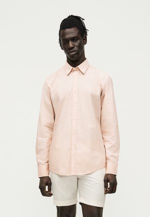 Mand med fletninger iført en lyserød langærmet skjorte med knapper og beige knælange shorts foran en ensfarvet baggrund.