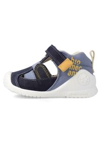 Sandalia de tela en azul marino y celeste con suela de goma blanca, cinta de velcro, detalles recortados y un parche de acento amarillo.