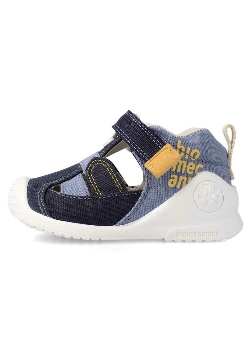 Sandalia de tela en azul marino y celeste con suela de goma blanca, cinta de velcro, detalles recortados y un parche de acento amarillo.