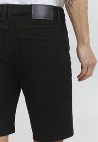 Șorturi negre din denim, cu o etichetă din piele pe spate, buzunare laterale și buzunare pe spate, având un design ajustat și un tiv curat. Detaliu texturat al țesăturii vizibil.