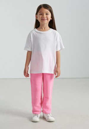 Maglietta bianca a maniche corte con una sfumatura rosa, abbinata a pantaloni da tuta rosa chiaro. Completa l'outfit un paio di sneakers bianche.