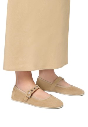 Beige suede flats with a rounded toe, adjustable strap featuring metal eyelets, and a lightweight white sole. Smooth texture throughout.