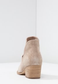 Bottine beige en daim avec talon carré en bois empilé et détail découpé sur le côté, posée sur une surface blanche unie.