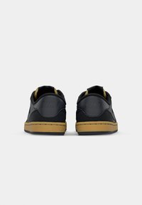 Ténis pretos de couro com detalhes em camurça, sola de borracha plana e sola exterior castanha. Apresenta o logótipo da Nike no calcanhar. Design arredondado.