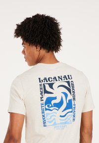 Kremo spalvos marškinėliai su mėlyna ir balta grafiką ant nugaros, kuri vaizduoja bangas ir abstrakčius formas su tekstu "LACANAU."
