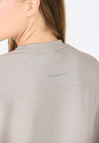 Chemise à manches courtes, de couleur beige clair avec une texture lisse, présentant un détail de couture subtil et le texte "ENDURANCE" imprimé en bleu clair au dos.