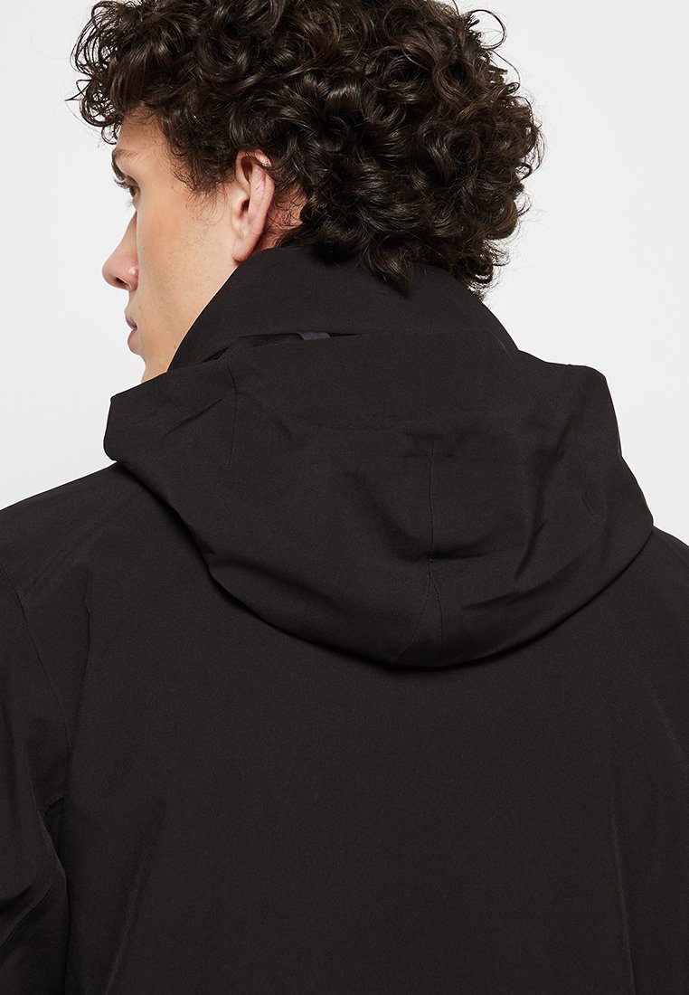 Giacca nera impermeabile con cappuccio aderente, texture liscia e dettagli di cucitura discreti. Il design è minimalista e funzionale.