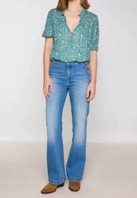 Blusa verde con motivo floreale e maniche corte, abbinata a jeans flare azzurri. Stivali alla caviglia marroni visibili in basso.