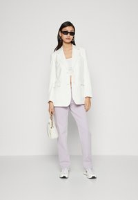 Blazer bianco a righe sottili sopra un crop top bianco, pantaloni lavanda, occhiali da sole neri e una borsa bianca fluffy. Le sneakers completano l'outfit.