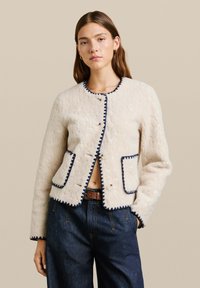 Witte fleecejas met golvende navyblauwe rand, ronde halslijn, twee voorkruispockets en knoopsluiting. Textielstof met een zachte uitstraling.