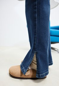 Džínsy tmavomodrej farby s bočnými rozparkami na lemoch a hnedou semišovou obuvou na sklz s detailom pracky.