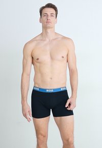 Black boxer briefs with a blue elastic waistband featuring the "BOSS" logo. They have a snug fit and smooth texture.