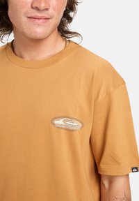 Joven con piel clara y pecas, vistiendo una camiseta de Quiksilver de color mostaza con el logo en el pecho y la manga.