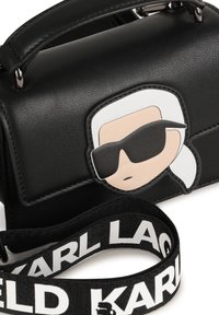 Borsa a tracolla in ecopelle nera con patch a forma di faccia di cartone animato, raffigurante una figura con capelli bianchi e occhiali da sole. Include una tracolla nera con logo.