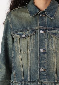Giubbotto in denim blu sbiadito con chiusura a bottoni, due tasche sul petto e collo. Presenta cuciture visibili e una finitura texturizzata.