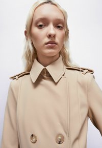 Béžový dvojradový kabát s vysokým golierom, ramennými vycpávkami a hladkou textúrou. Má veľké, priehľadné gombíky.