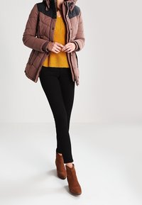 Brown quilted jacket with a black shoulder panel, worn over a yellow knitted sweater and black leggings, paired with brown heeled boots.