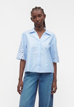 Femme aux cheveux tressés portant un chemisier rayé bleu clair avec des manches à découpes florales et un jean bleu ample, debout devant un fond blanc.