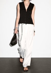 Svart ärmlös topp med djup V-ringning och knappstängning, kombinerad med vita lösa byxor och svarta sandaler. Håller en svart handväska.