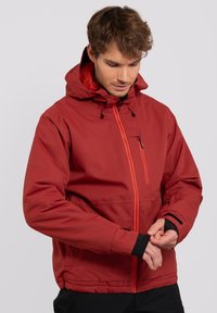 Chaqueta impermeable roja con capucha, que cuenta con un brillante zipper naranja, puños negros y un bolsillo en el pecho. Cuello forrado de forro polar para mayor calidez.