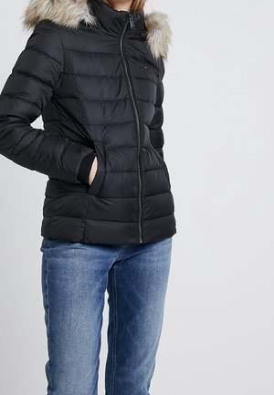 Person wearing a black quilted jacket with a fur-trimmed hood and hands in pockets, paired with blue denim jeans.