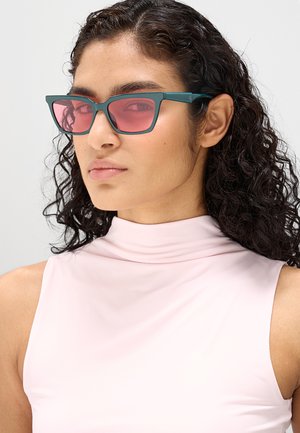 Donna con capelli ricci scuri, che indossa occhiali con montatura turchese e lenti rosa e un top senza maniche rosa pallido a collo alto.