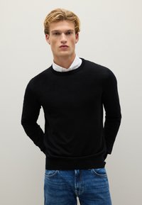 Schwarzer Rundhalsausschnitt-Pullover aus weichem Strickstoff, mit langen Ärmeln und ribbeligem Saum, kombiniert mit einem weißen Hemd mit Kragen darunter.