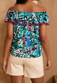 Top décolleté avec un imprimé animalier vibrant vert et rose, présentant des bords froncés avec des pompons roses. Tissu léger, coupe ample.