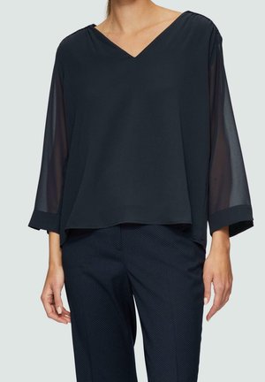 Femme portant un chemisier noir ample à col en V avec des manches trois-quarts transparentes et un pantalon bleu marine foncé avec un motif à pois subtil.