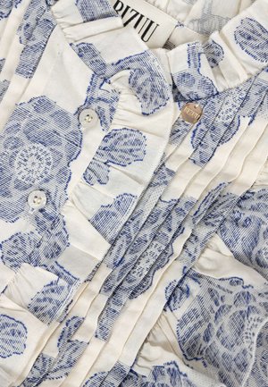 Biały materiał z niebieskim fakturowanym kwiatowym nadrukiem, pionowymi plisami, białymi guzikami, falbankami na krawędziach oraz małą okrągłą metalową metką z nazwą marki.